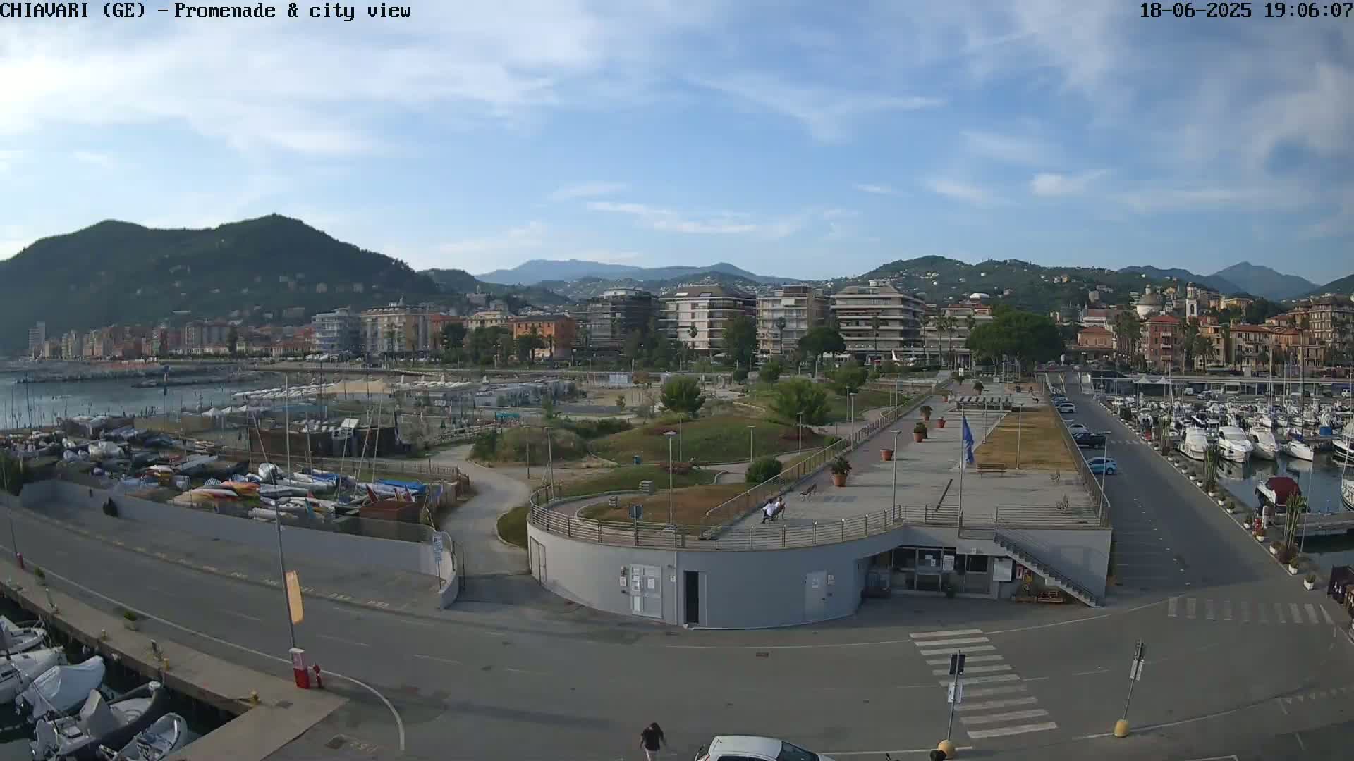 A waterfront promenade with a marina filled with boats, a city skyline backed by mountains, and a mostly sunny sky.