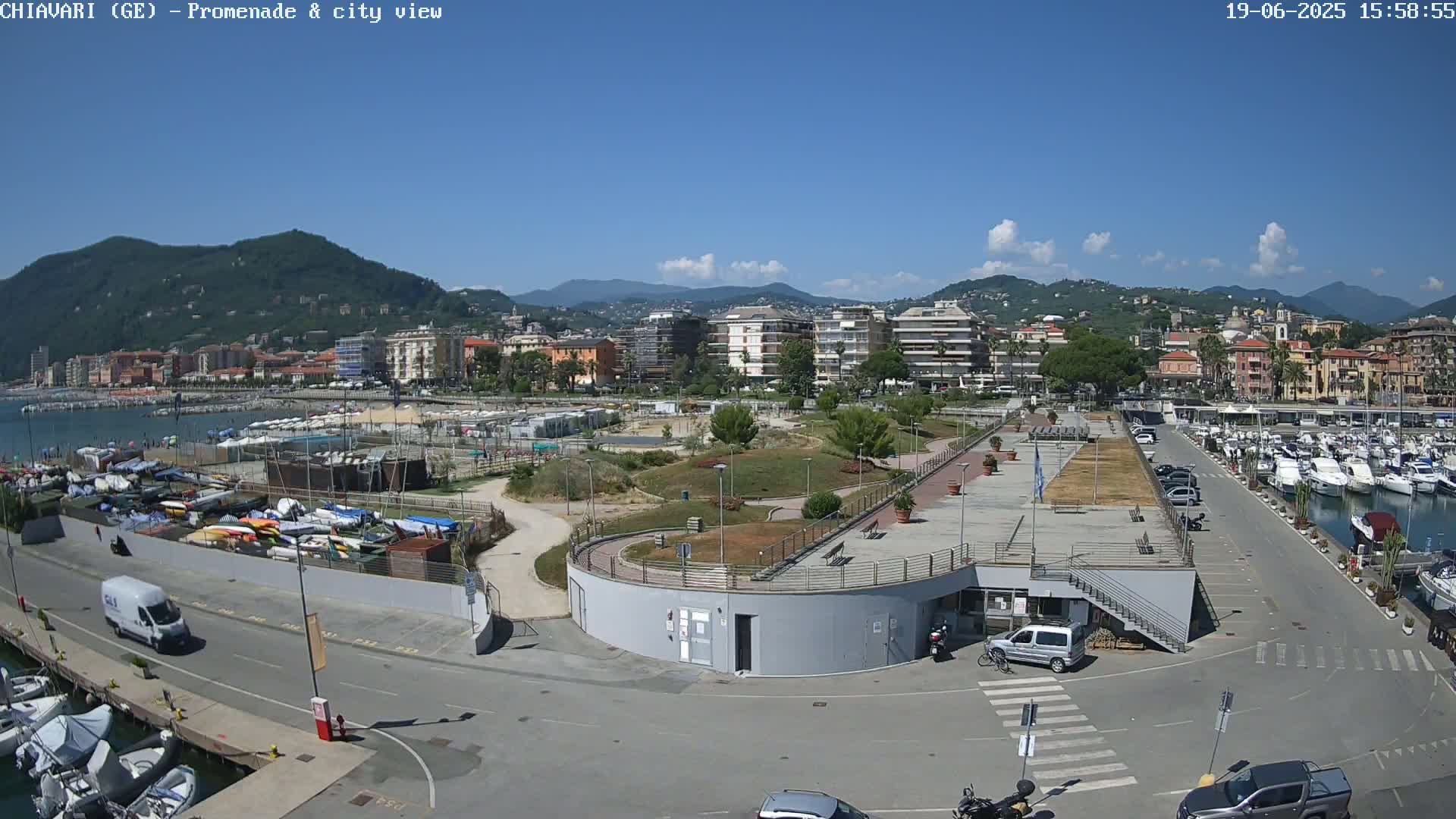 A sunny day overlooking a harbor filled with boats, a promenade, and a coastal town nestled against mountains.