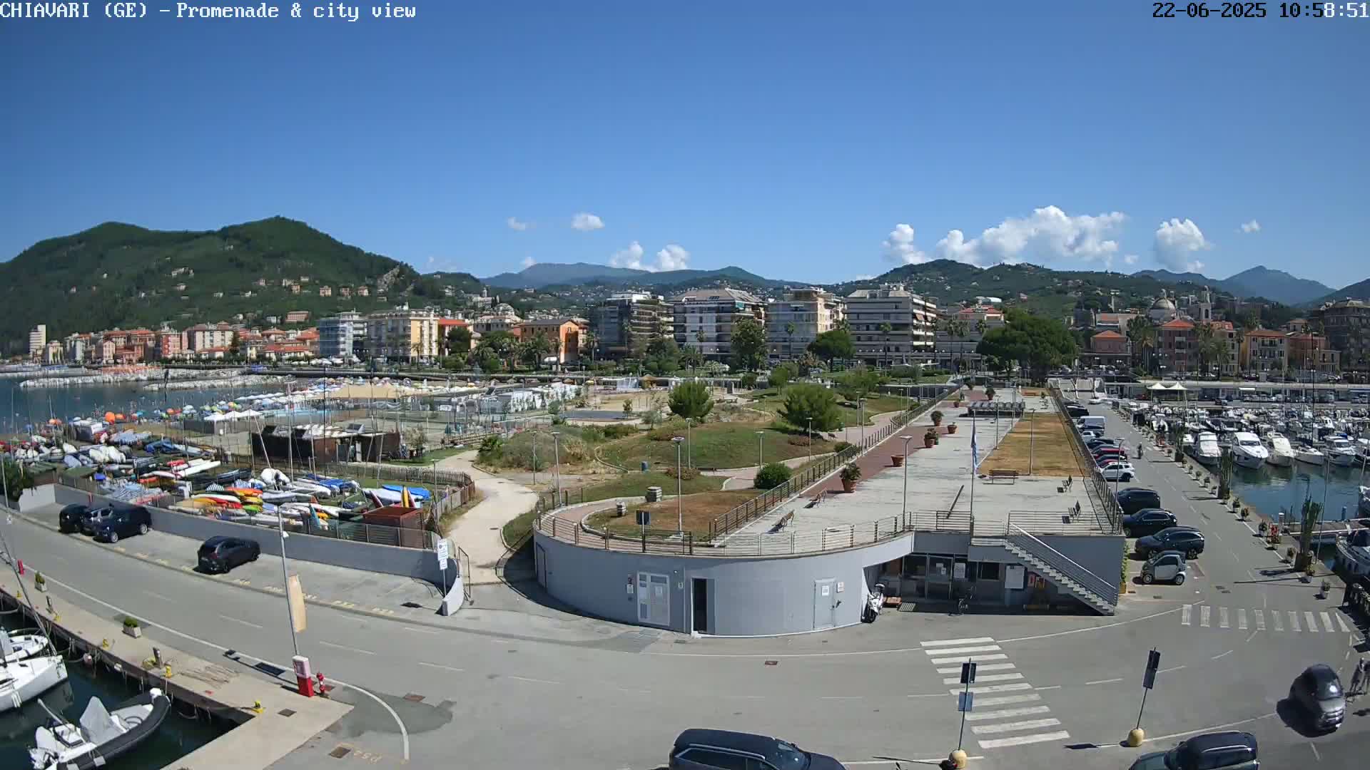 A seaside town under a mostly sunny sky is seen from an elevated viewpoint, showing a marina with numerous boats, a promenade, a park, and buildings along the coast.