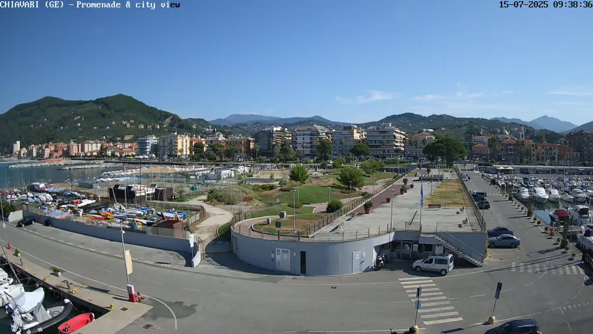 A sunny waterfront scene shows a marina filled with boats, a paved promenade, and a town nestled against hills under a clear blue sky.