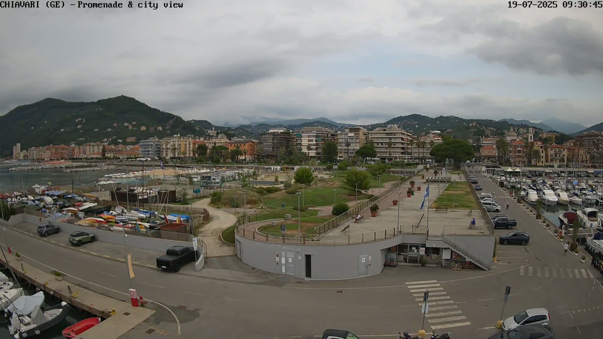 A waterfront promenade and marina with a city and mountains in the background under a cloudy sky.