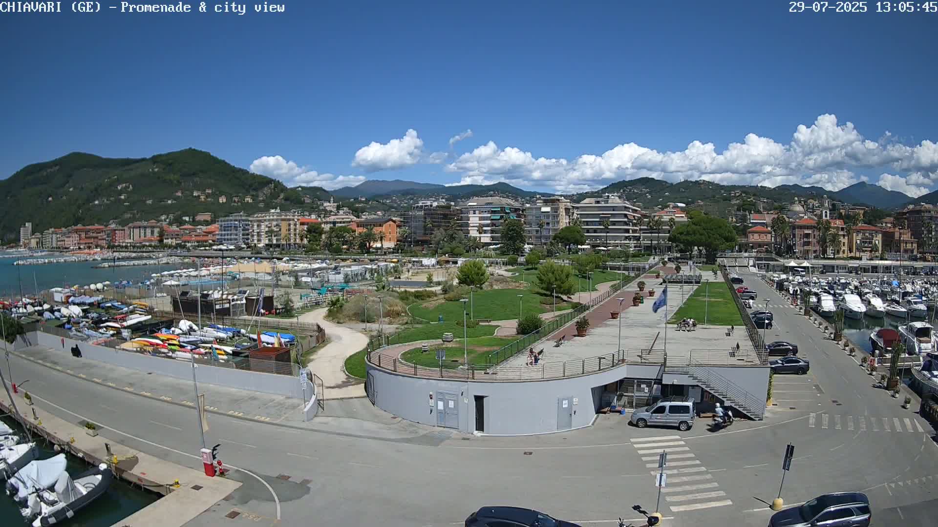 A waterfront promenade and marina with a city and mountains in the background under a cloudy sky.