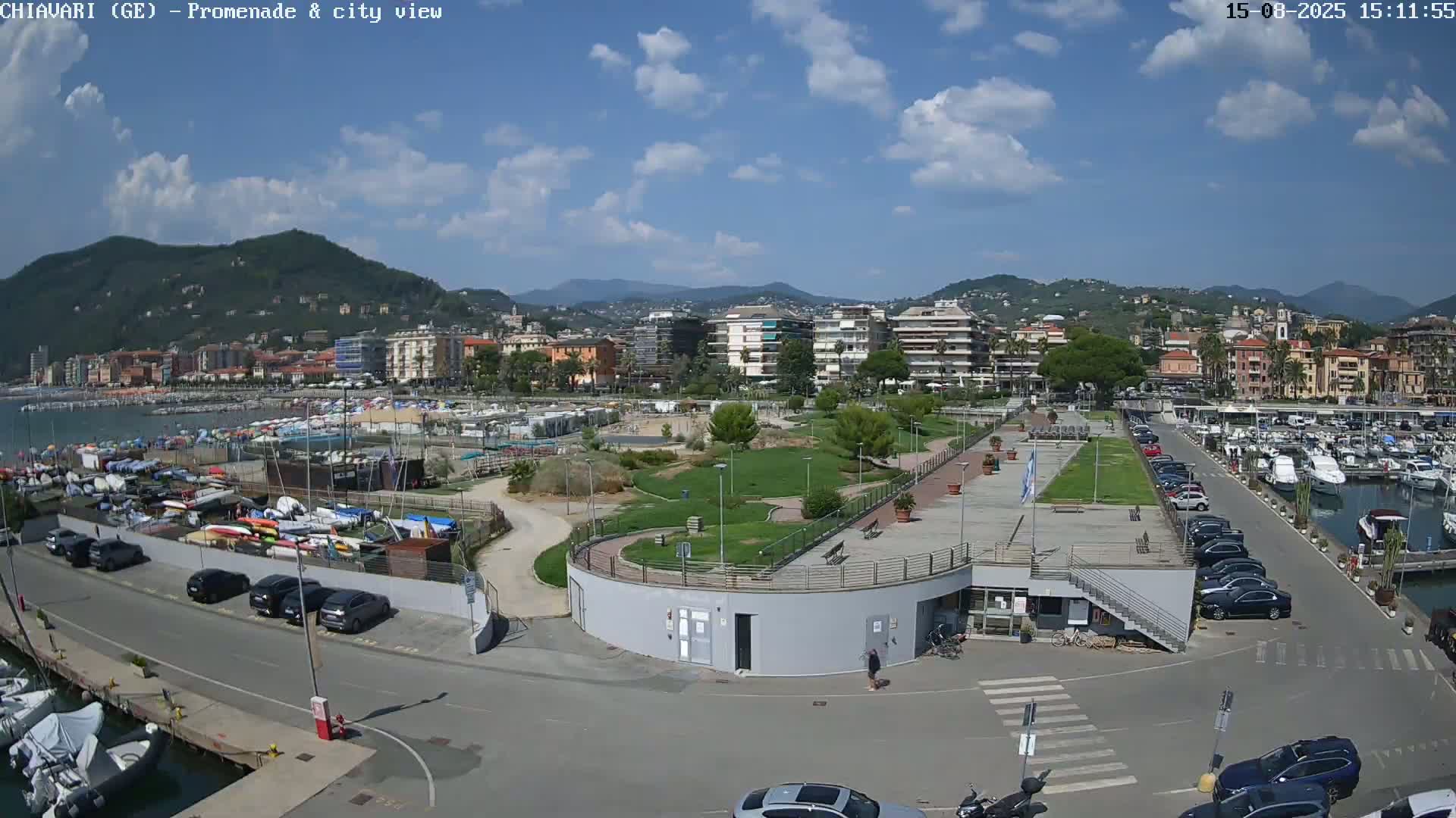 An aerial view shows a harbor filled with boats, a grassy promenade, and a town nestled against a mountain range under a clear blue sky.