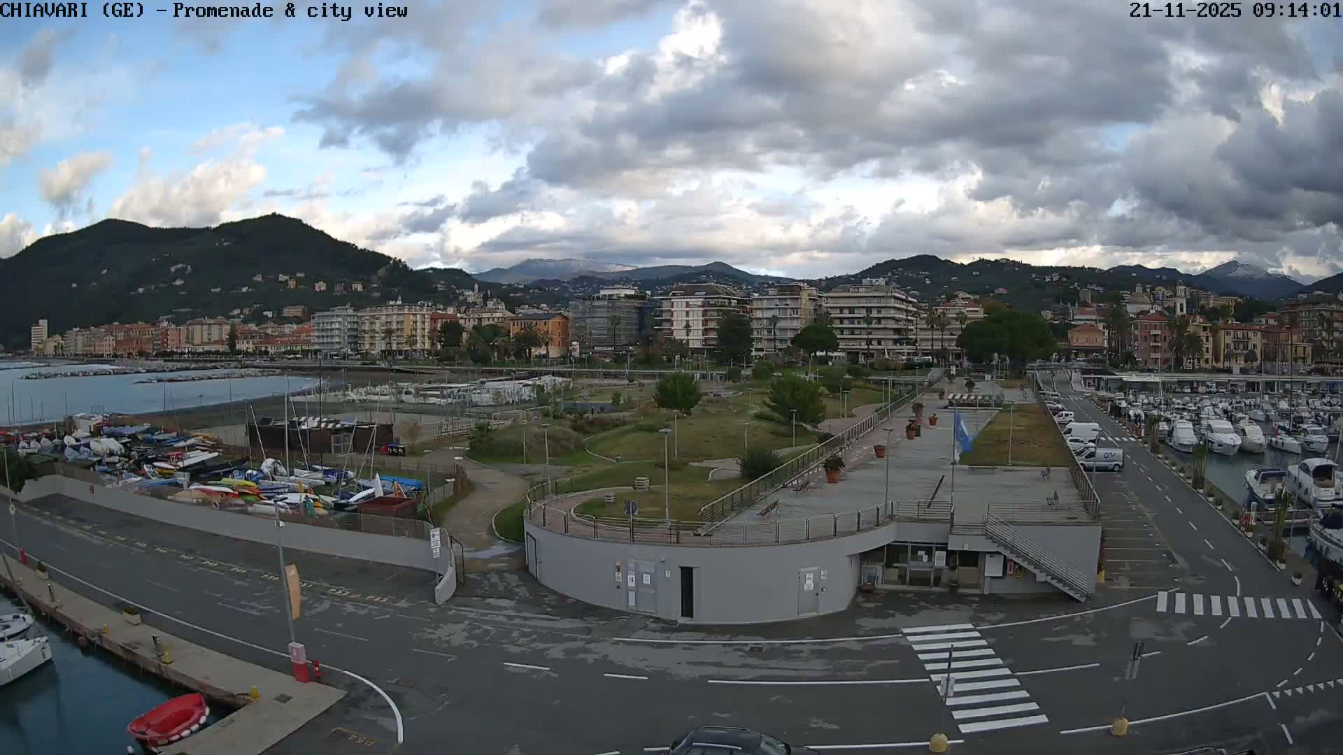A bustling coastal scene is depicted under a partly cloudy sky, showing a marina packed with yachts and boats on both land and water, a modern promenade, and a town nestled against dark hills with distant snow-capped mountains.
