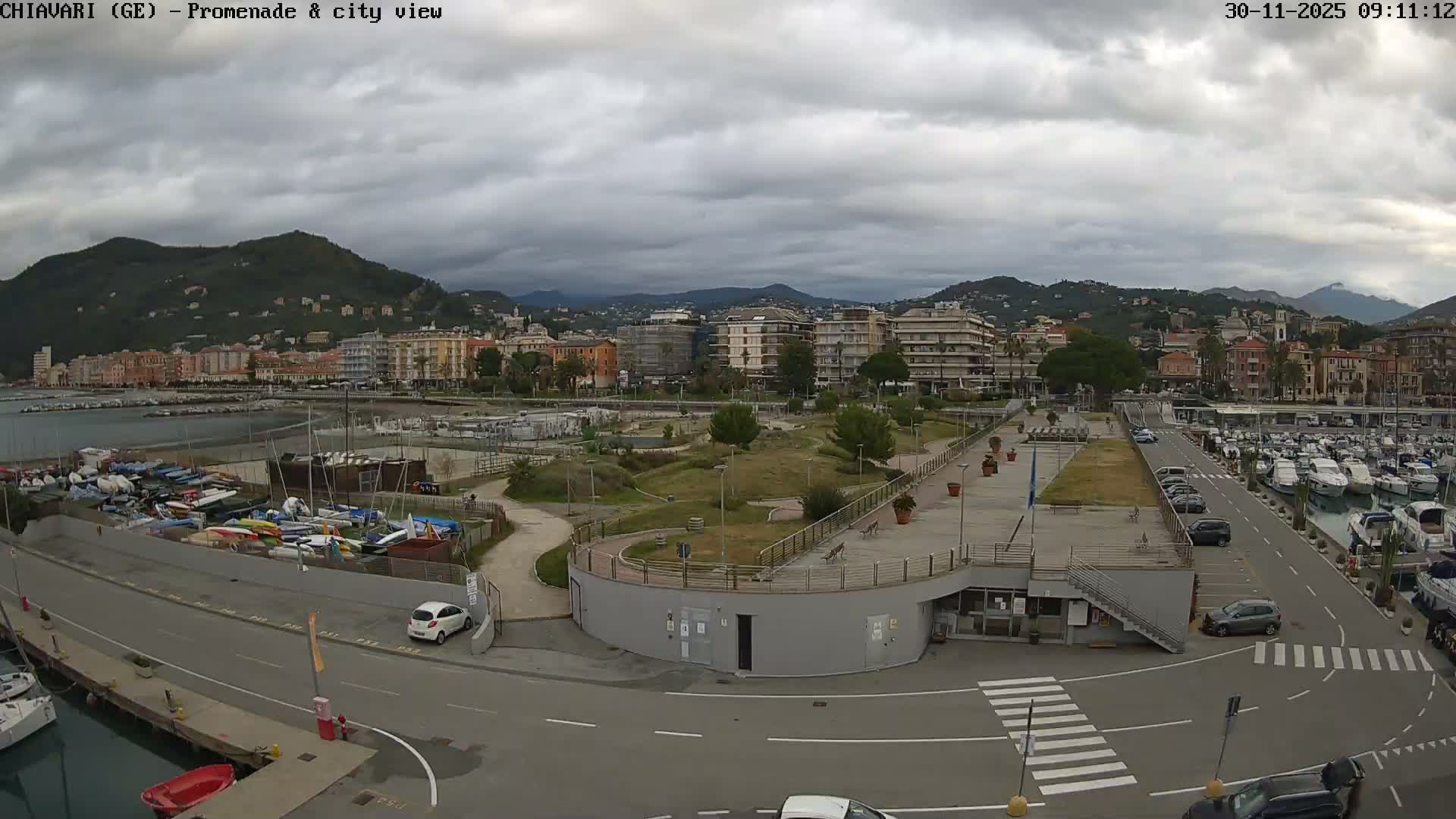 A wide view captures a coastal city's harbor filled with various boats, a busy road, a modern promenade, and buildings nestled against lush mountains under a heavily overcast sky.