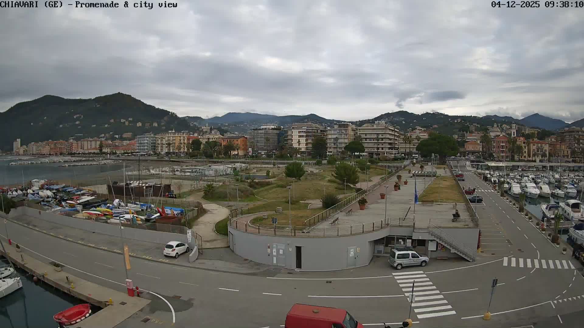 An overcast day reveals a panoramic view of a vibrant coastal city featuring a marina teeming with boats, a dedicated area for stacked small watercraft, a green park, and numerous buildings lining the base of a range of lush mountains.
