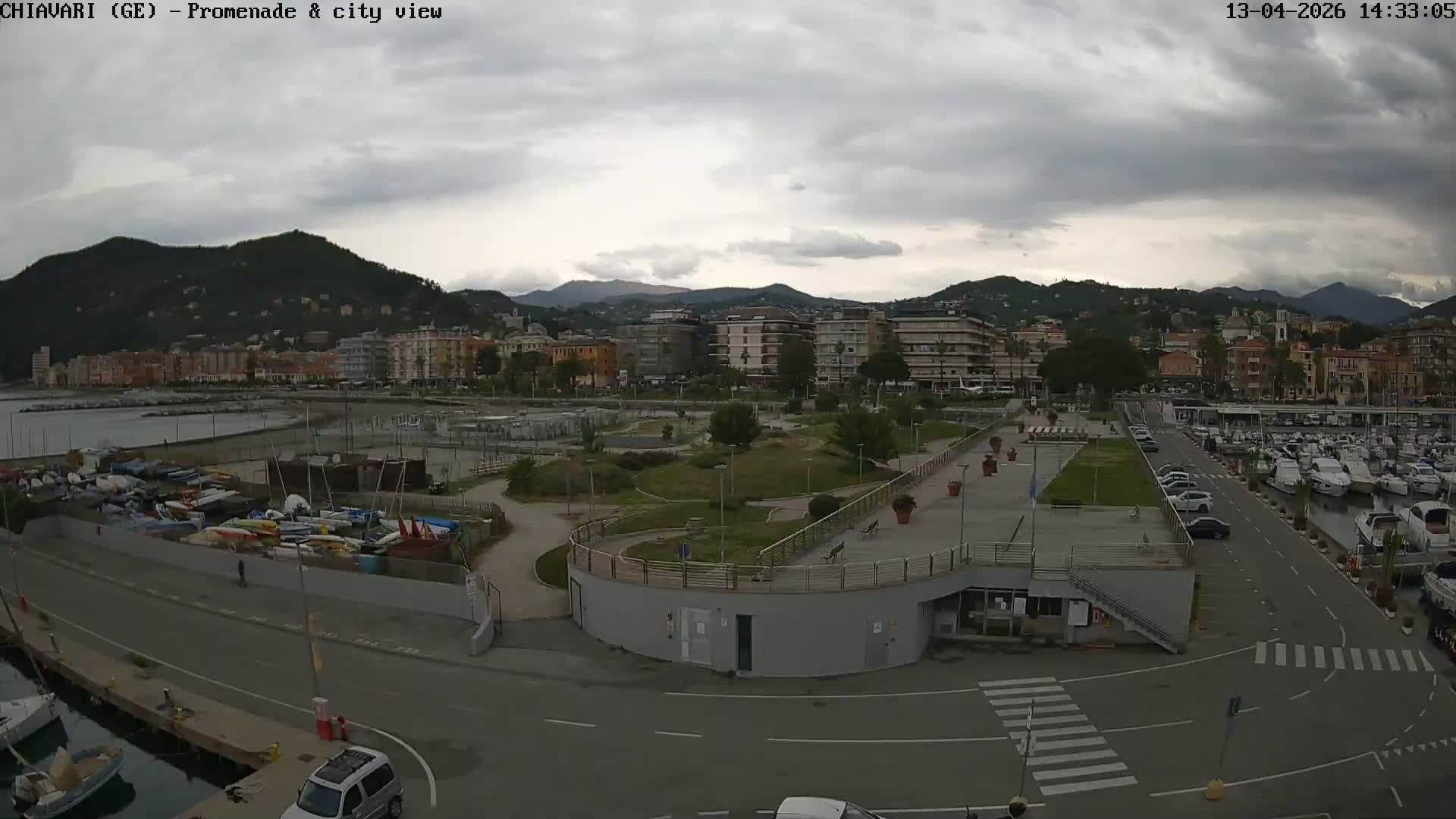 An overcast day reveals a panoramic view of a vibrant coastal city featuring a marina teeming with boats, a dedicated area for stacked small watercraft, a green park, and numerous buildings lining the base of a range of lush mountains.