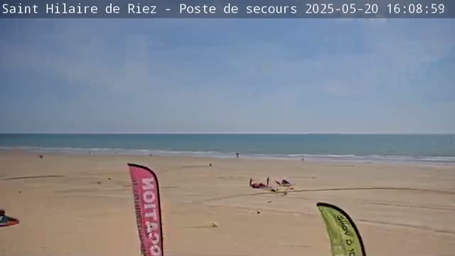 A sandy beach with calm ocean waves under a clear blue sky shows a few people and some kitesurfing equipment on the sand.