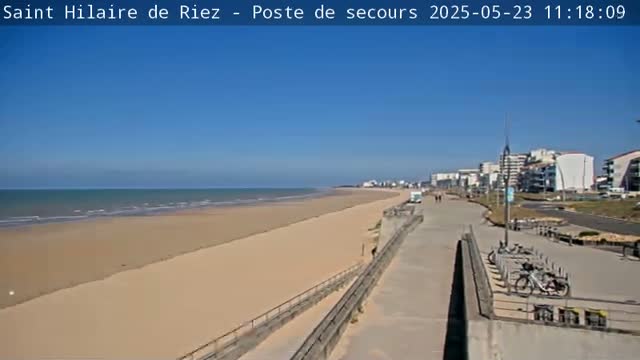 A wide sandy beach stretches to a calm ocean under a clear blue sky, with a boardwalk and buildings visible in the distance.