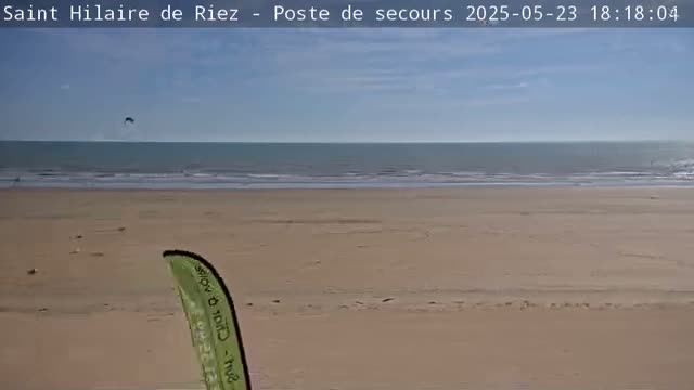 A sandy beach with a calm ocean under a mostly clear sky, and a kitesurfer visible in the distance.