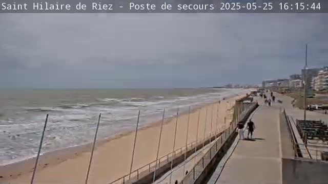 A sandy beach with moderate waves under an overcast sky is bordered by a paved walkway with a few pedestrians, and buildings are visible in the distance.