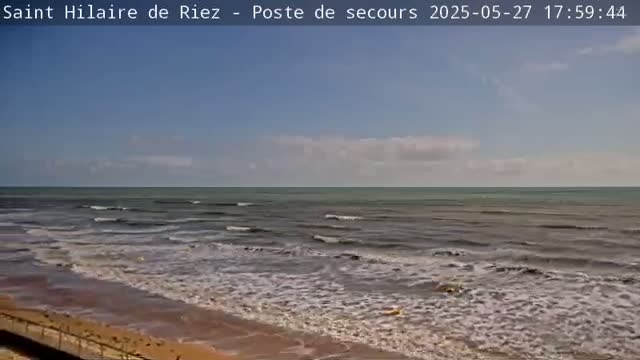 A mostly sunny day shows a gently wavy ocean meeting a sandy beach.