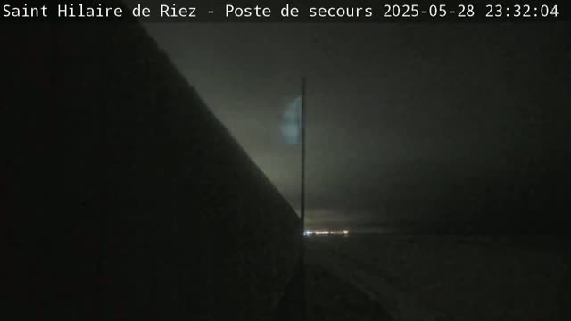 A dimly lit, nighttime scene shows a flagpole with a faintly visible flag, a dark wall or structure to the left, and a hazy, light-colored area in the distance.