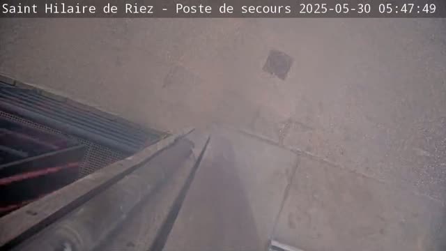A hazy view from a high angle shows a long, dark structure that appears to be a metal pole or pipe, with a concrete or paved area visible below, alongside some metallic grating and pipes.