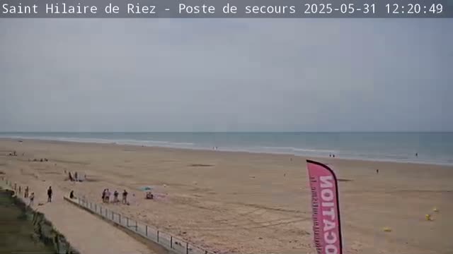 A mostly overcast day at a wide sandy beach with calm ocean waves and a few people scattered across the sand.