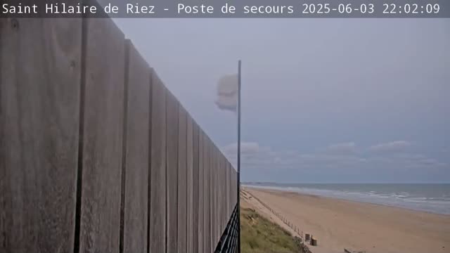 A tall wooden fence overlooks a wide sandy beach and a calm ocean under an overcast sky.