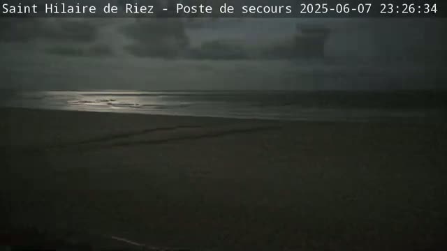 A dark, overcast night scene shows a calm ocean meeting a wide, dark sandy beach.