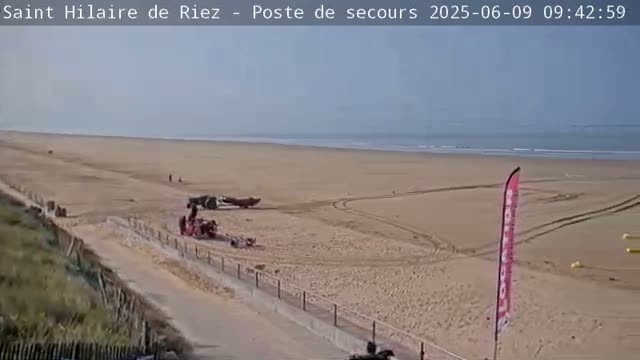 A sandy beach with calm ocean waves under a clear sky shows several vehicles and people near a paved path leading from a grassy dune.