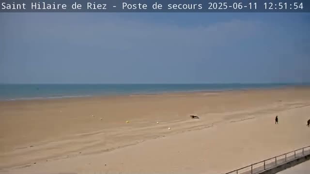 A wide sandy beach stretches to a calm ocean under a mostly clear sky, with a few people walking near the water's edge.