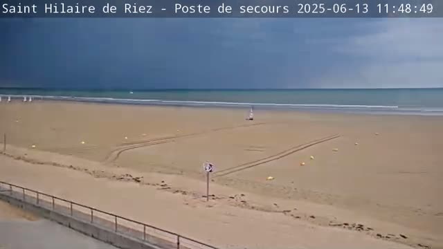 A mostly cloudy sky overlooks a wide sandy beach with tire tracks and a few small objects scattered across it, and a calm ocean in the background.