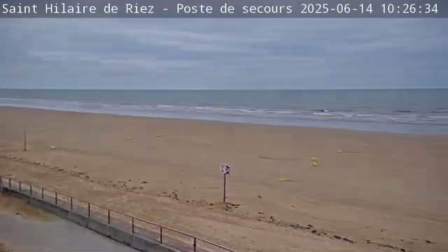 A mostly-clear, overcast day shows a wide, sandy beach sloping gently down to a calm ocean with small waves.