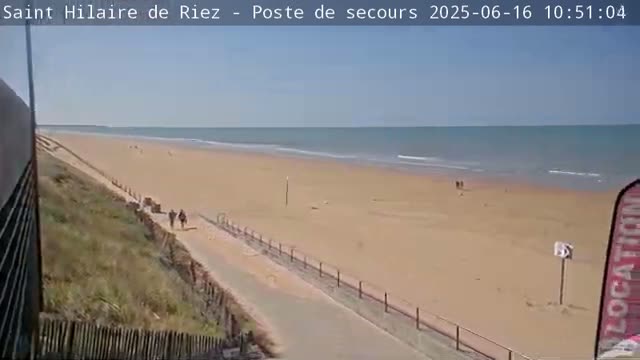 A sandy beach with a calm ocean under a clear sky is viewed from a dune, with a few people walking along a path.