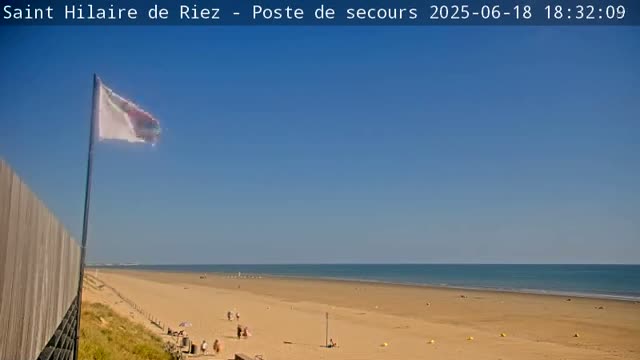 A sandy beach stretches to a calm ocean under a clear blue sky, with a flagpole and a few people visible.