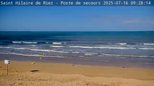 A sandy beach slopes down to a calm ocean with small waves under a clear blue sky.