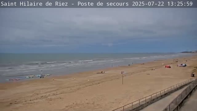 A sandy beach with a few people on it stretches along a calm ocean under a cloudy sky.