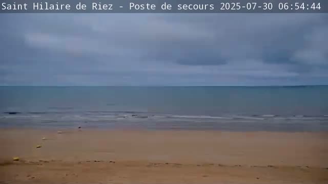 A sandy beach with a few people on it stretches along a calm ocean under a cloudy sky.