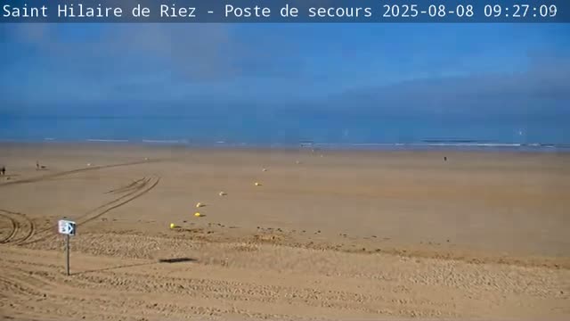 A mostly clear, blue sky overlooks a wide, sandy beach with a calm ocean in the distance, and a few people are visible in the far distance.