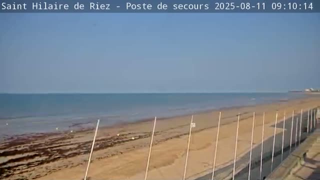 A mostly clear, sunny day reveals a wide sandy beach and calm ocean, with several tall poles lining the beach.