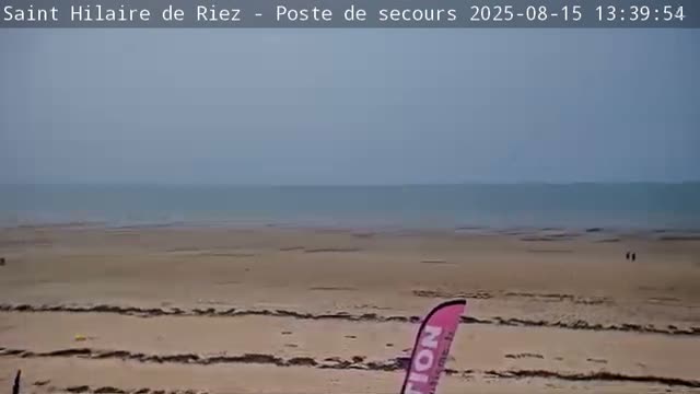 A mostly clear, sunny day reveals a wide sandy beach and calm ocean, with several tall poles lining the beach.