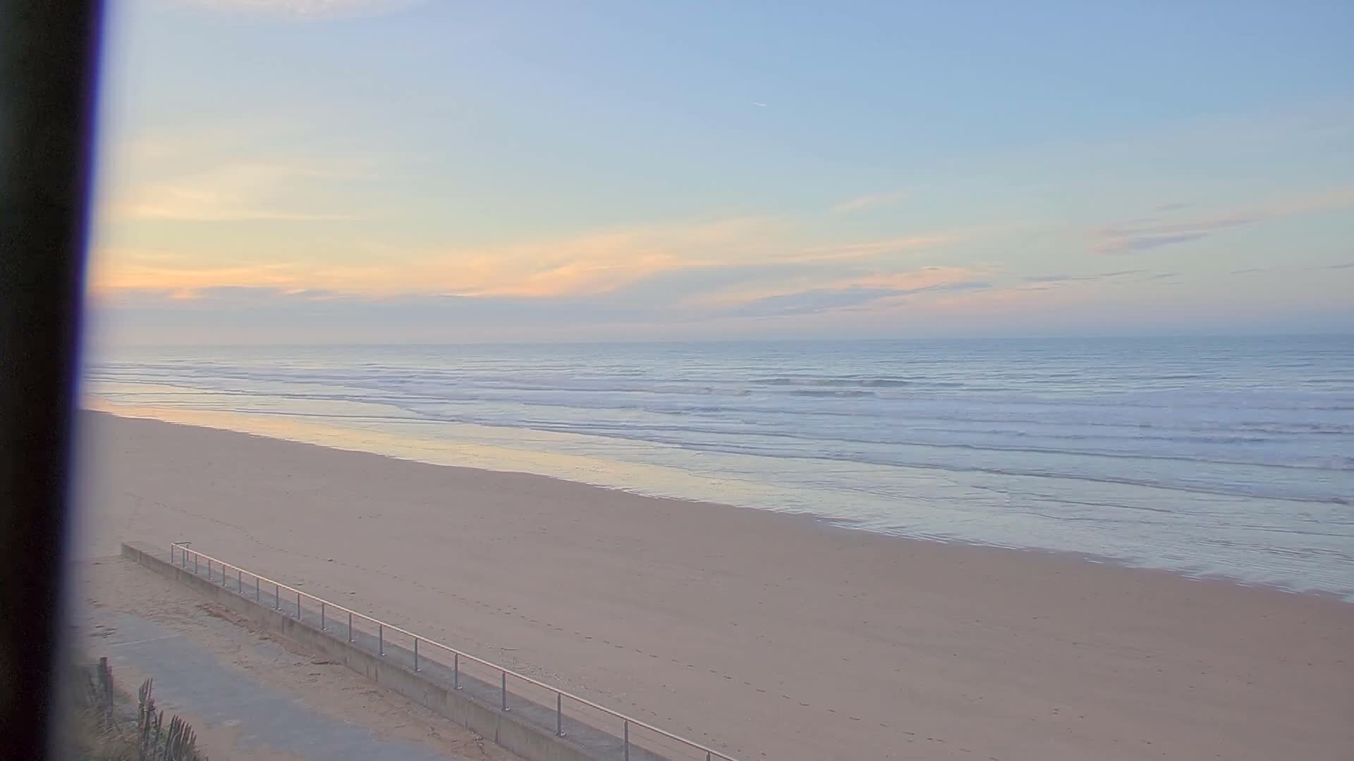 A wide sandy beach with gentle waves meets a calm ocean under a clear, pastel sky with scattered soft clouds, and a concrete walkway with a railing is visible along the lower left, indicating fair weather.