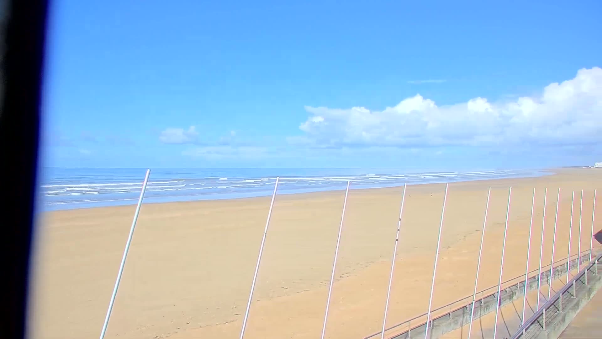 A wide sandy beach with gentle waves meets a calm ocean under a clear, pastel sky with scattered soft clouds, and a concrete walkway with a railing is visible along the lower left, indicating fair weather.