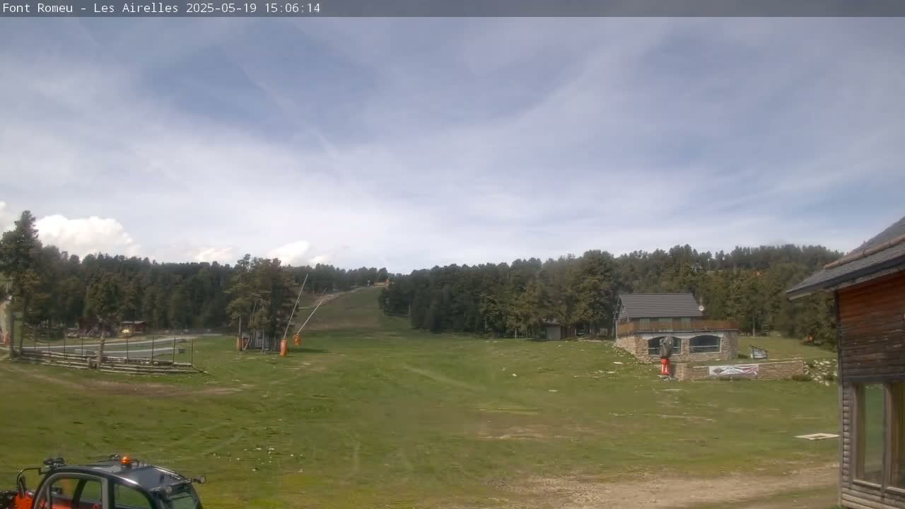 A mostly clear sky overlooks a grassy ski slope with a few buildings and a sparsely wooded hillside in the background.
