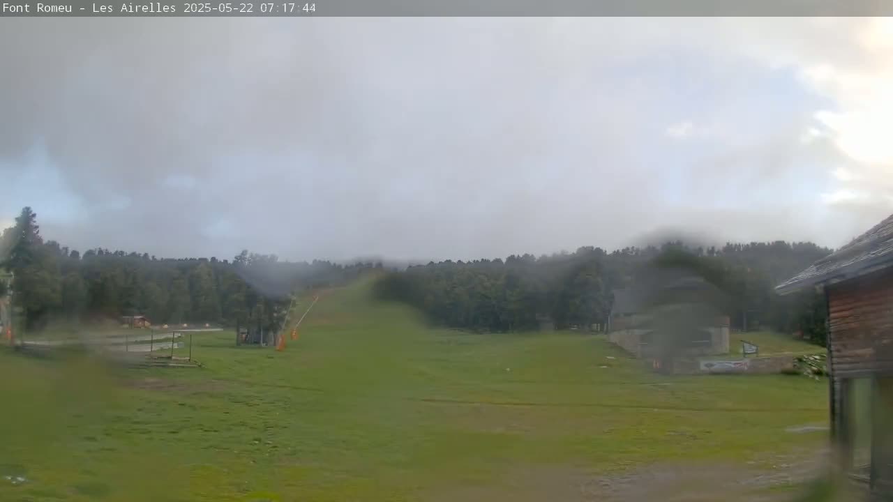 A misty, rainy scene shows a grassy field with a ski lift in the distance, bordered by a dark forest and a wooden building in the foreground.