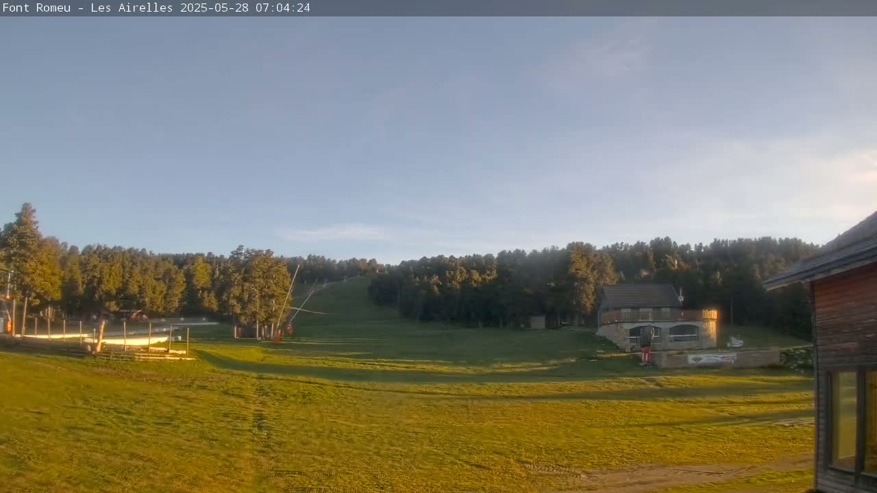 A grassy field slopes gently down toward a line of trees, with a small building and ski lift visible in the mid-ground under a clear, bright sky.