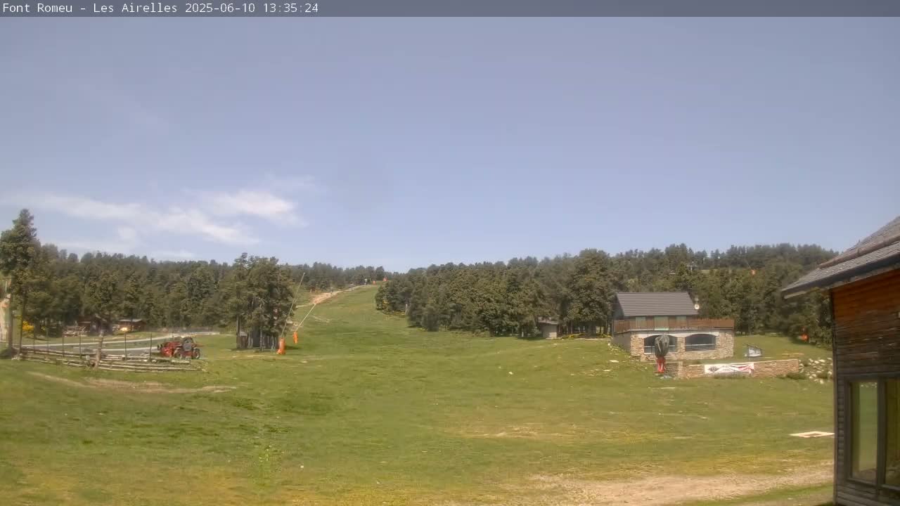 A mostly sunny day reveals a grassy hillside with a few buildings, a forest of evergreen trees, and a ski lift in the background.