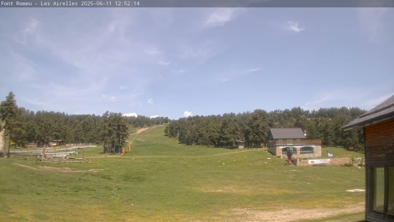 A grassy area with a ski lift and buildings sits in front of a forest under a mostly clear, bright sky.