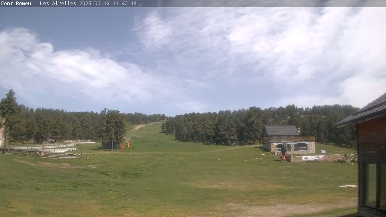 A partly cloudy sky overlooks a grassy field with a ski lift and several buildings nestled among a coniferous forest.