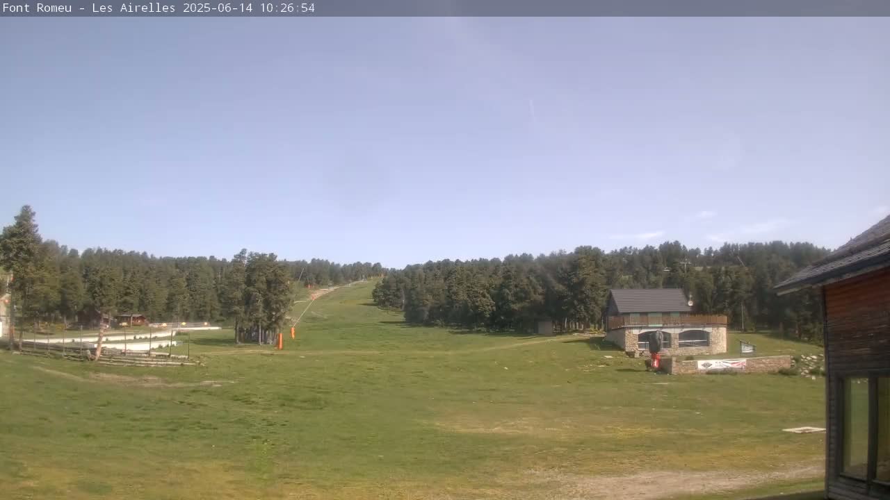 A grassy field and ski slope are visible under a clear sky, with a few buildings and a forest in the background.