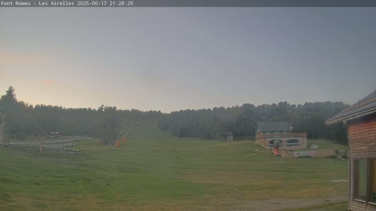 A grassy field with a line of trees in the background, a couple of buildings, and a ski lift under a hazy, light-grey sky.