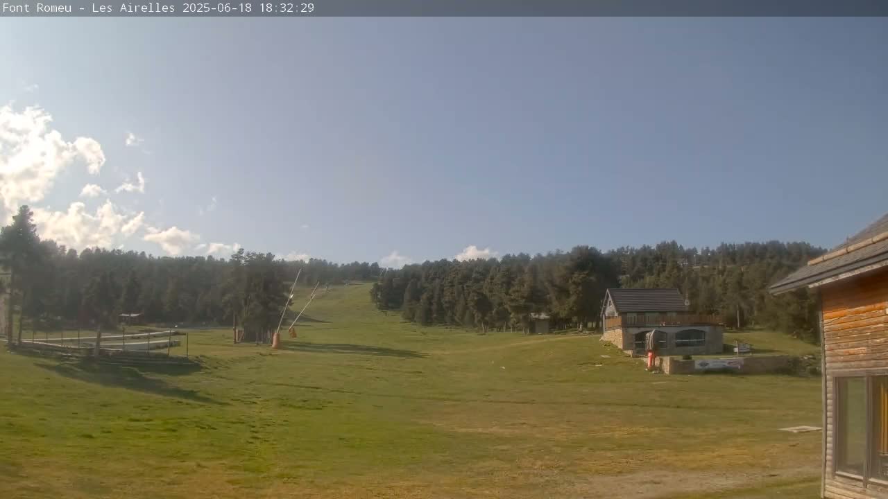A partly cloudy sky looks down on a grassy field, a small building, and a forested hillside with a ski lift in the distance.
