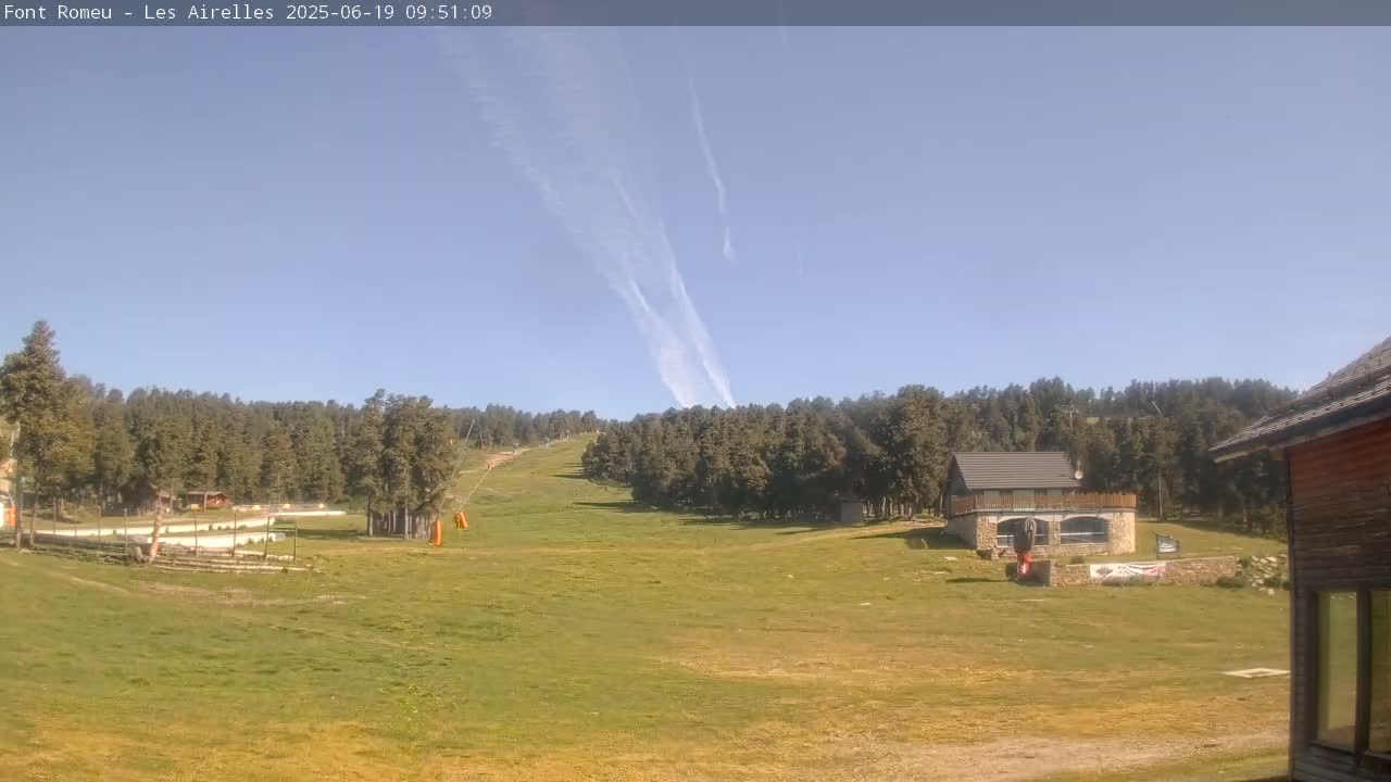 A grassy hillside slopes down towards a small building, with a forest of evergreen trees in the background under a mostly clear, blue sky with a few contrails.