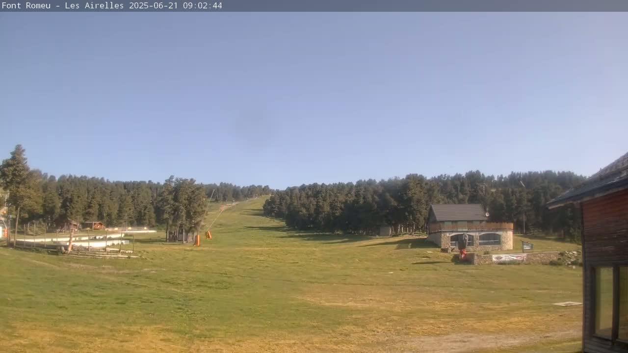 A grassy field and ski slope are visible under a clear blue sky, with a forest of evergreen trees in the background and several small buildings.