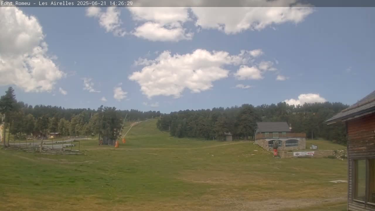 A partly cloudy sky overlooks a grassy field with a small building, trees, and a ski lift in the background.