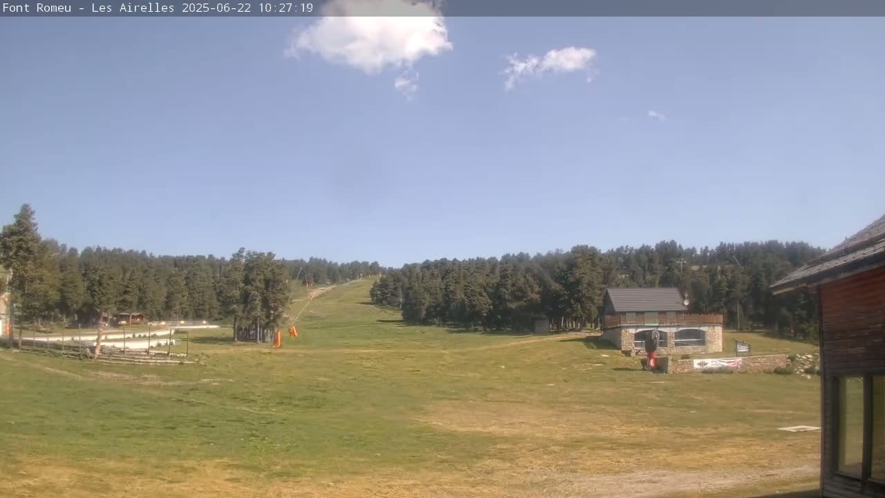 A grassy field with a small building and a ski lift in the background under a mostly sunny sky with a few scattered clouds.