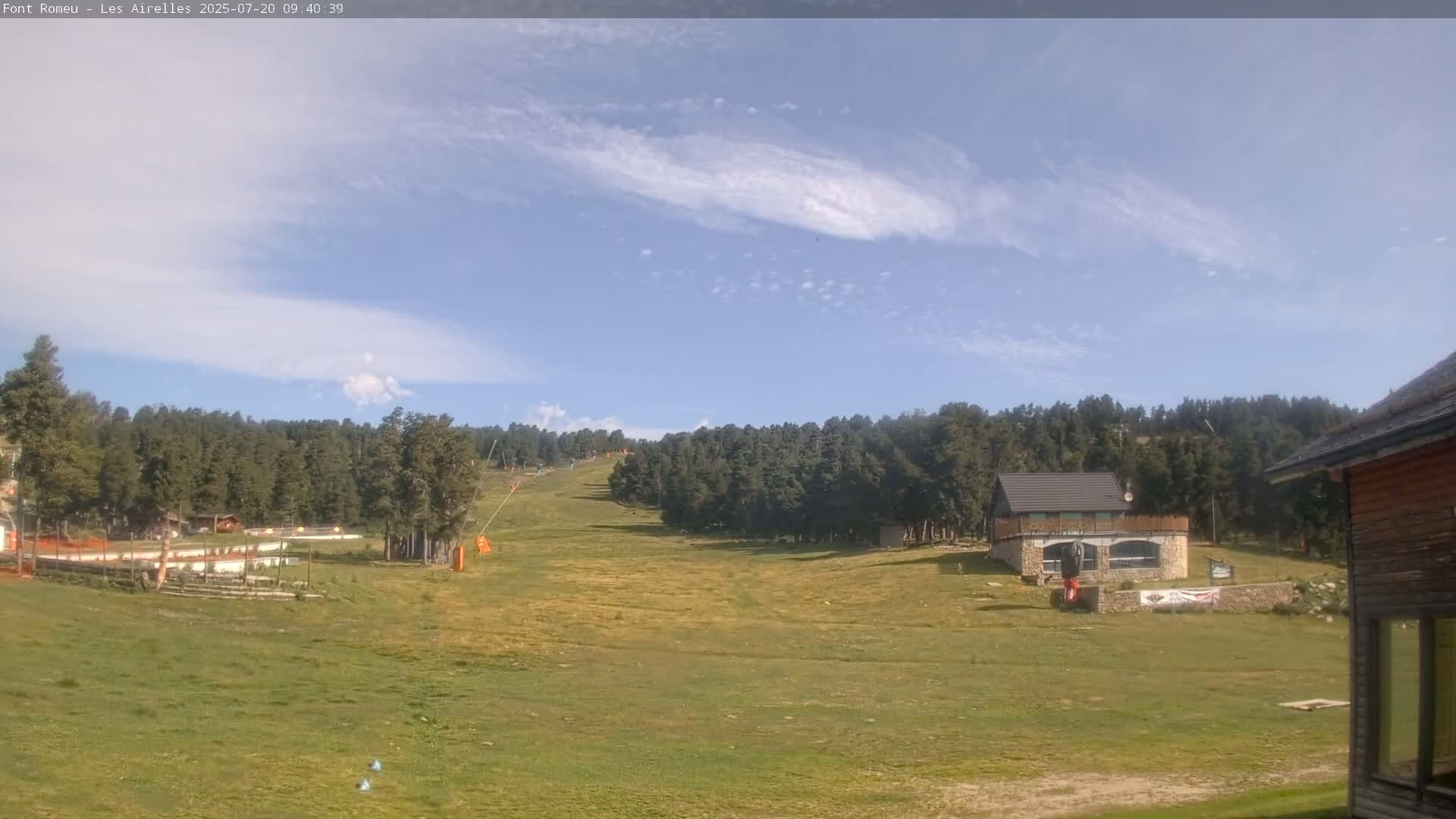 A grassy hillside with a ski lift and small buildings is visible under a mostly clear, bright blue sky with wispy clouds.