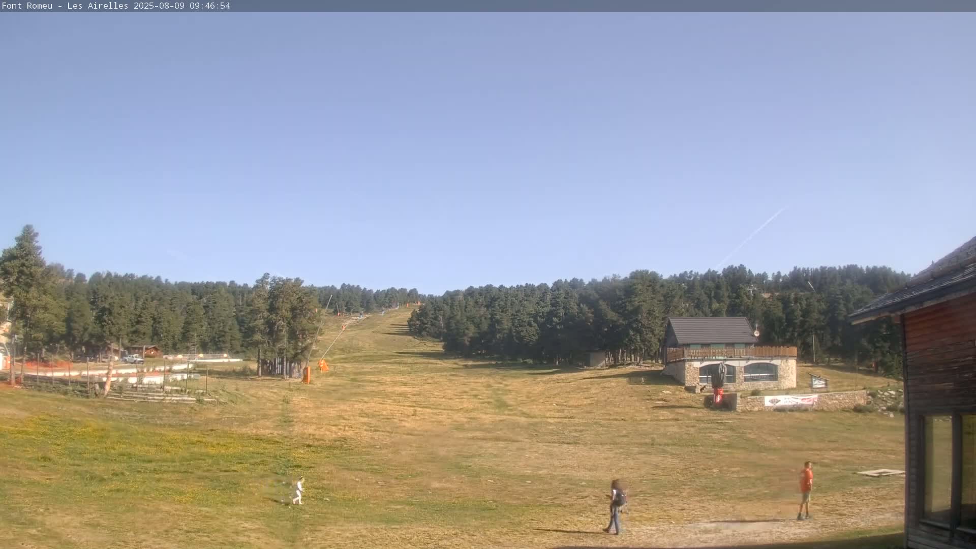 A sunny, clear day at a mountain resort shows a grassy area with a few people walking, a ski lift in the background, and some buildings.